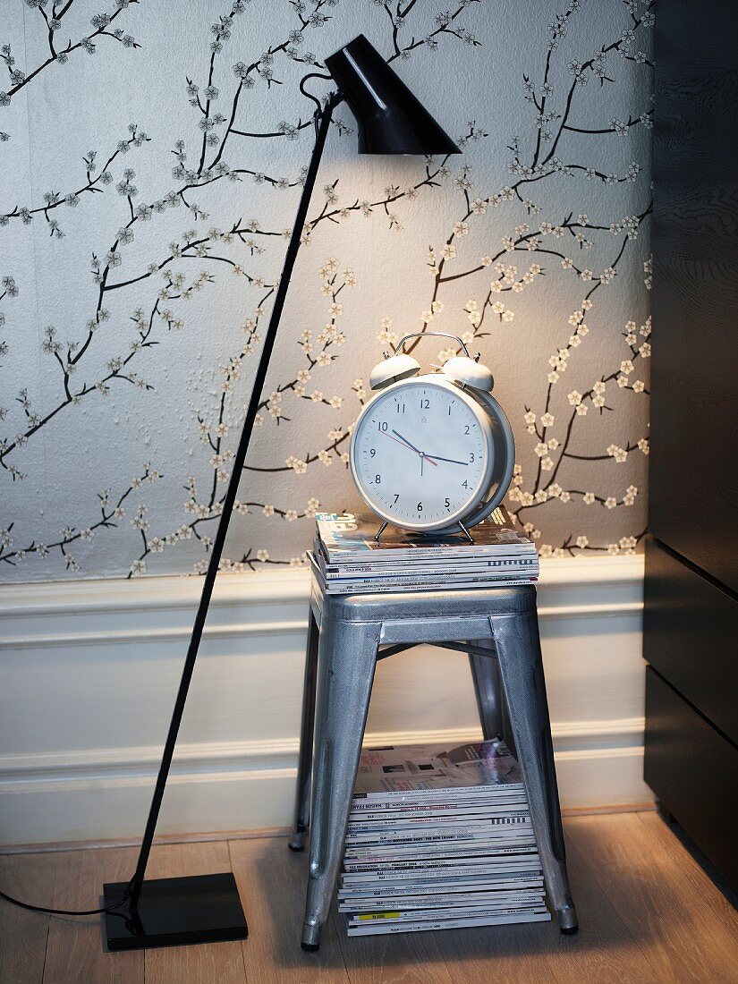 Newspapers and an alarm clock on a silver stool with a floor lamp next to it