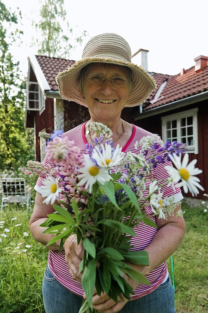 Ältere Frau mit Blumenstrauss im Garten vor Haus