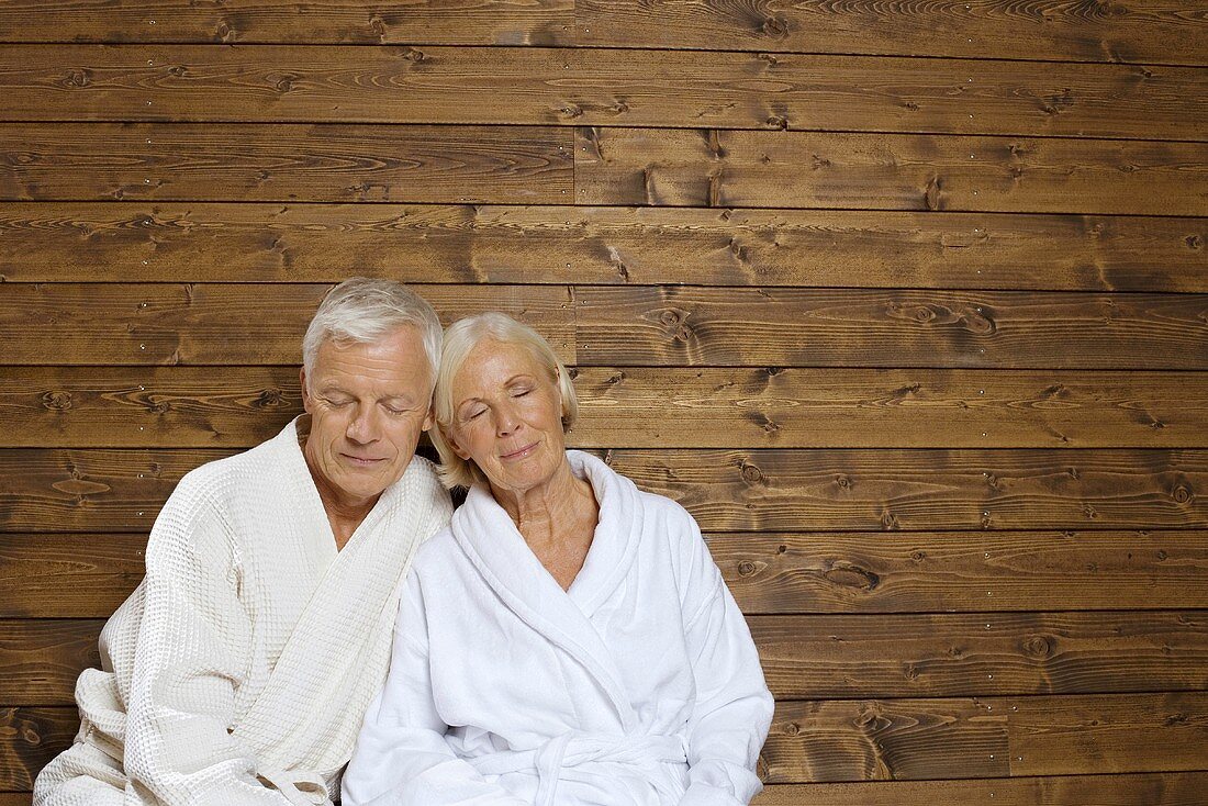 Älteres Paar in Bademänteln sitzt an Holzwand