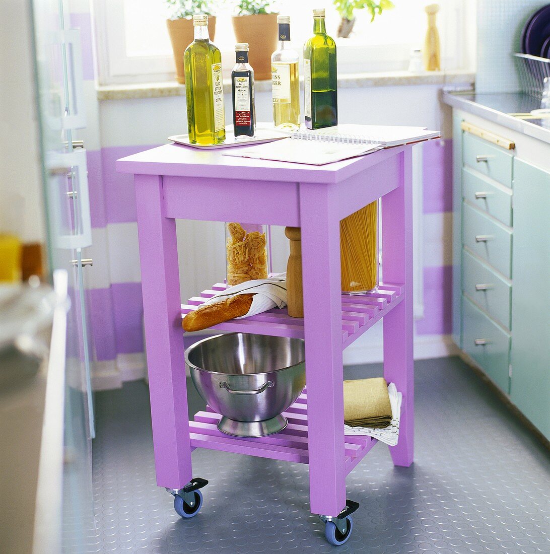 Pastellfarbener Küchenrollwagen mit Öl-und Essigflaschen, Gläsern mit Nudeln, einem Baguette und einer Edelstahlschüssel