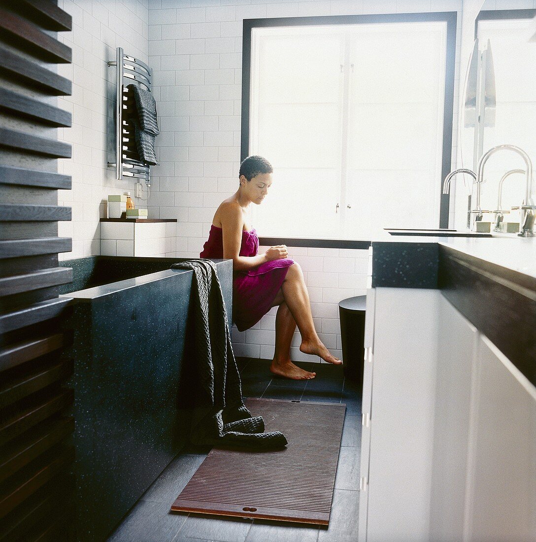 Junge Frau in ein violettes Badetuch gewickelt, in modernem Badezimmer sitzend