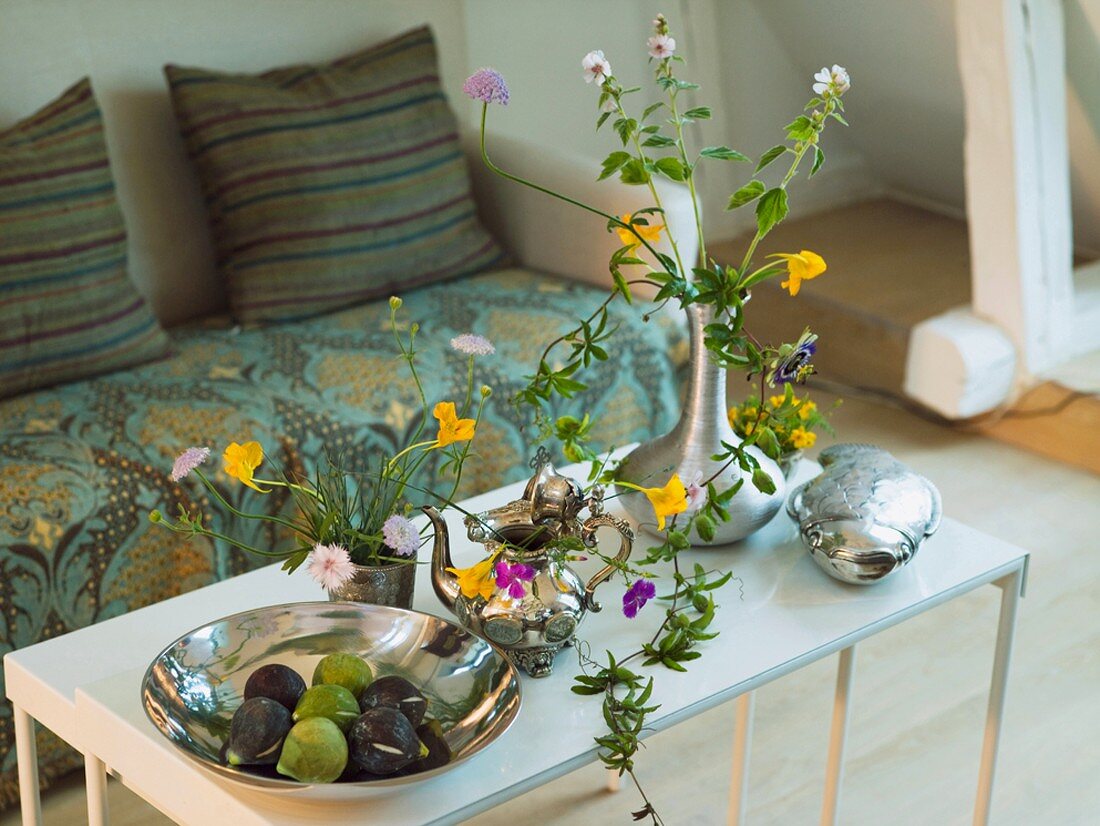Couchtisch mit silberner Obstschale mit Keramikfeigen, einer silbernen Kanne und Wiesenblumen in einer silbernen Vase