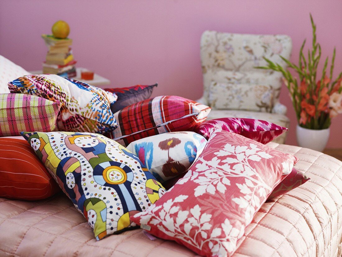 Verschiedene bunte Zierkissen auf Bett mit rosa Steppdecke; im Hintergrund ein Sessel, daneben eine Bodenvase mit großem Gladiolenstrauss