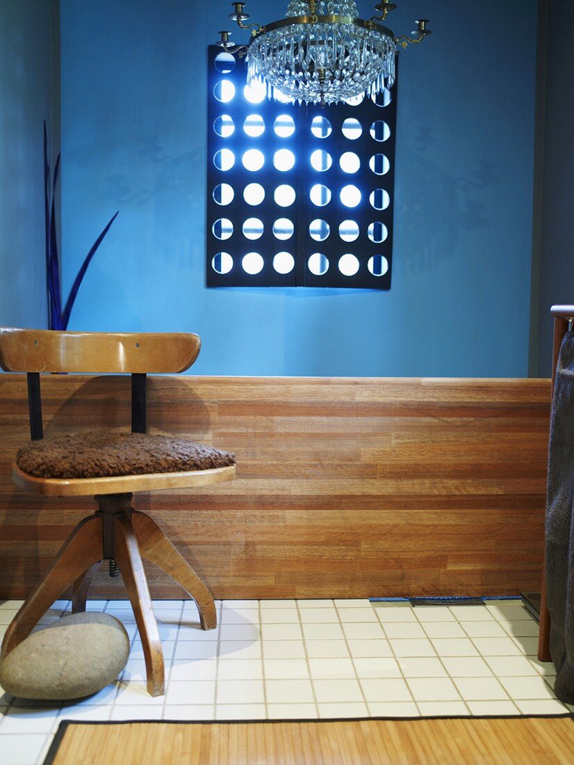 Drehstuhl aus Holz mit braunem Sitzpolster vor blauer Wand; darüber ein Kristallleuchter