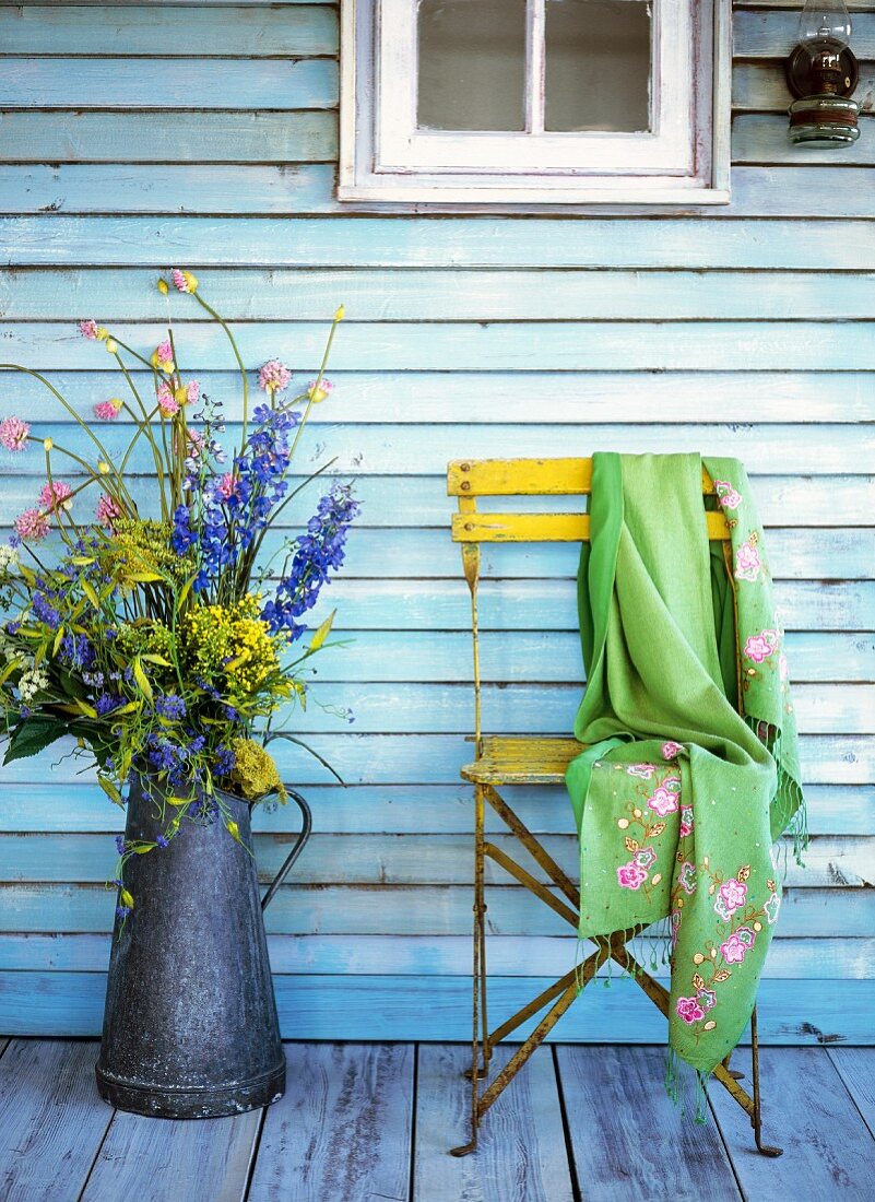 Meadow flowers in a zinc jug and an embroidered cashmere scarf on a garden chair against the wall of a house