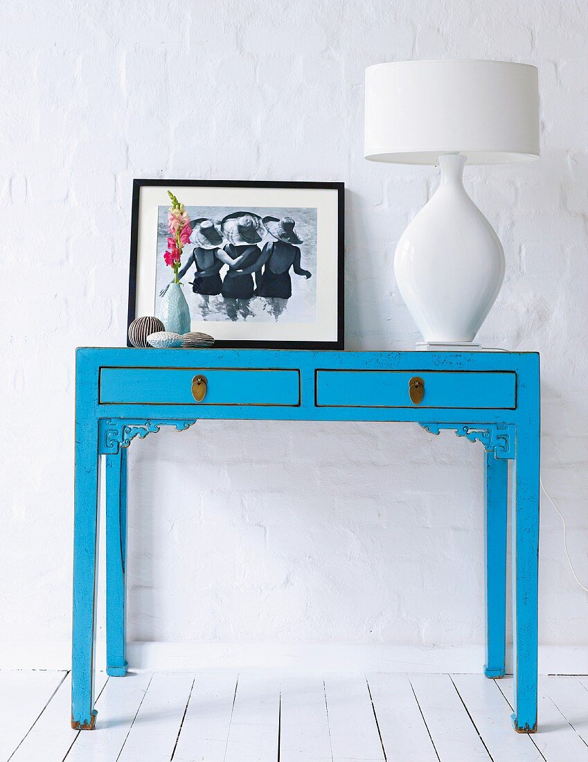Blauer Konsolentisch dekoriert mit weisser Tischleuchte & Fotographie vor weisser verputzter Steinwand