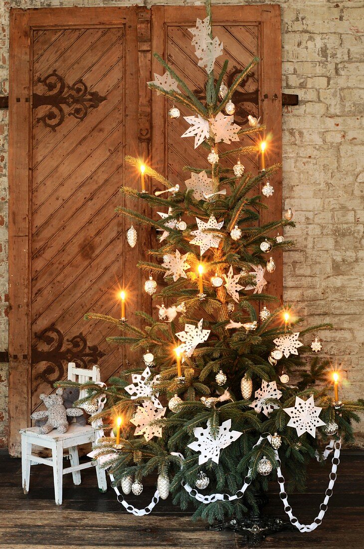 A Christmas tree in front of a rustic wooden door decorated with nostalgic paper stars and candles
