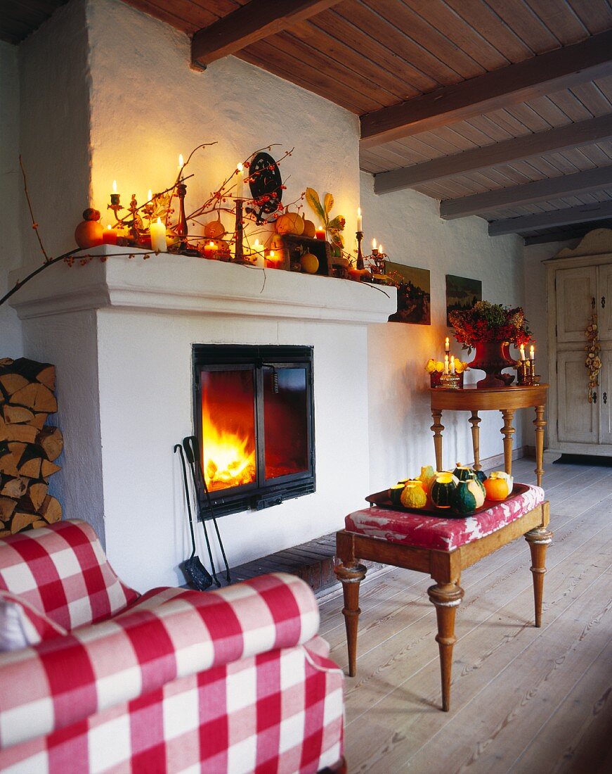 Herbstlich dekorierter, ländlicher Wohnraum mit Kaminfeuer, davor antiker, gepolsterter Schemel und Sessel mit kariertem Bezug