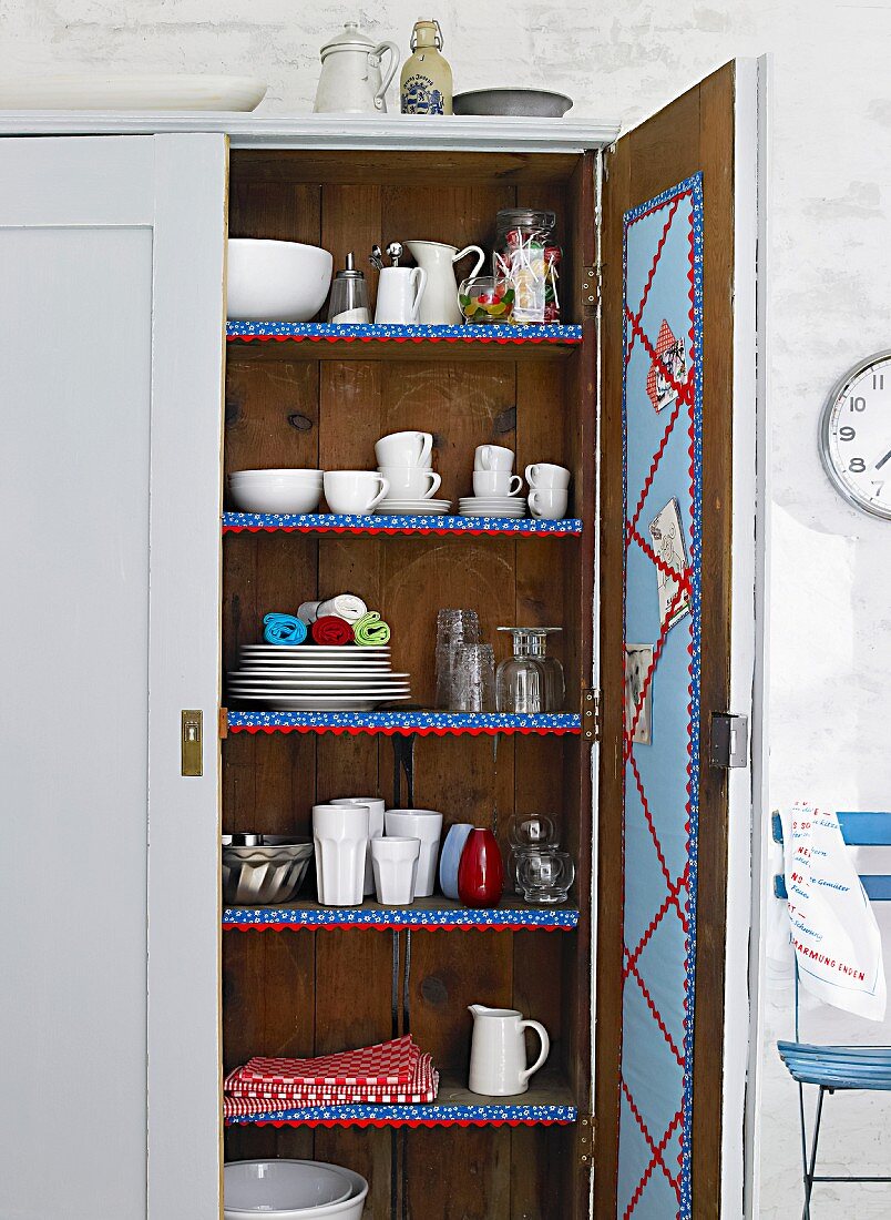 Geöffneter Küchenschrank aus Holz mit Geschirr auf Bordüren verzierten Regalen