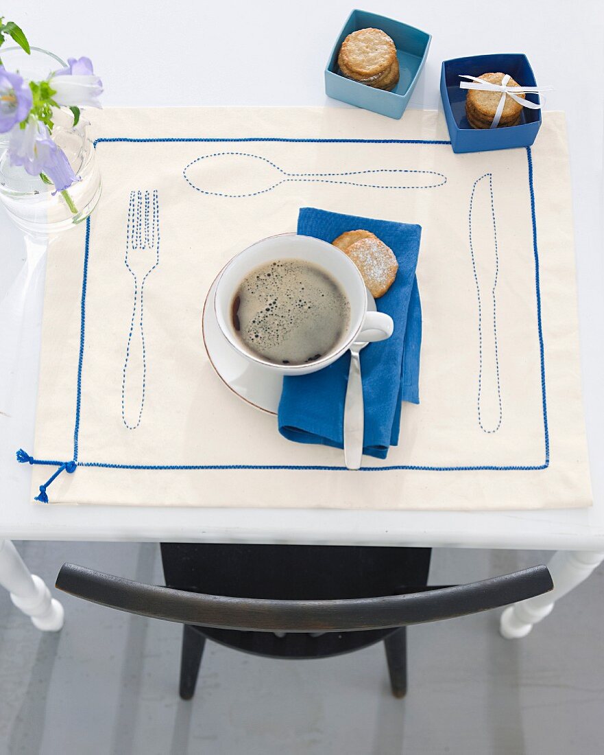 Kaffeetasse mit Gebäck auf dekorativem Platzset mit Besteckmotiv & Zierband