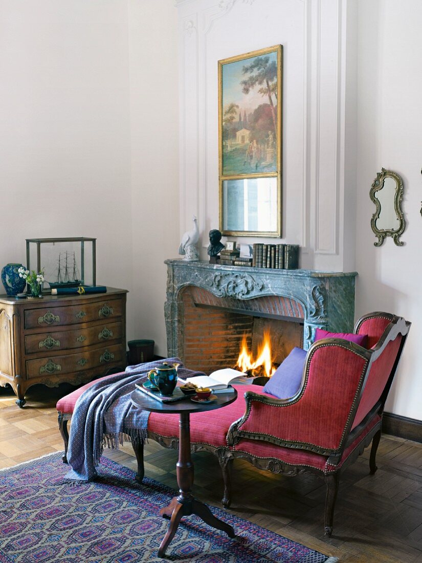 An antique chaise lounge with a red velvet cover and a side table next to an open fire