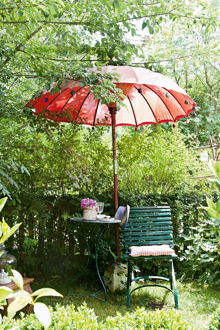 Sitzplatz in sommerlichen Garten mit Gartenstuhl, Gartentisch & rotem thailändischen Sonnenschirm