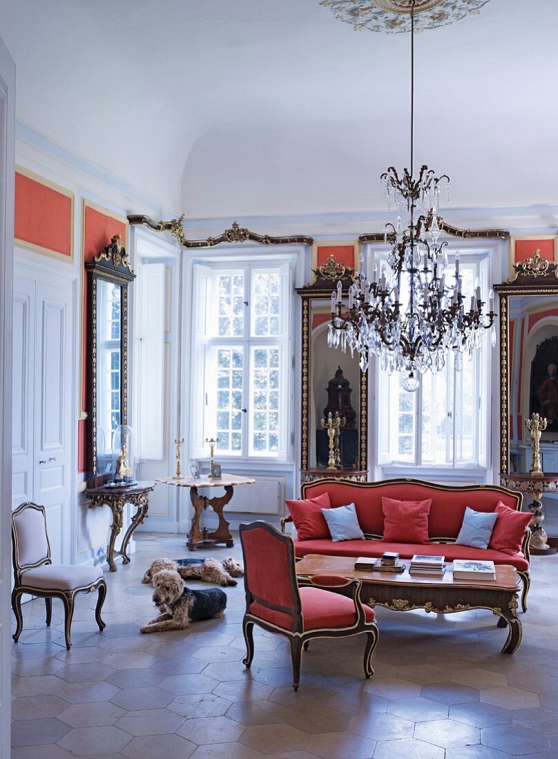 A grand living room Castle Dennenlohe with a Rococo armchair and matching sofa with a chandelier above it and dogs sitting on the honeycomb-tiled floor