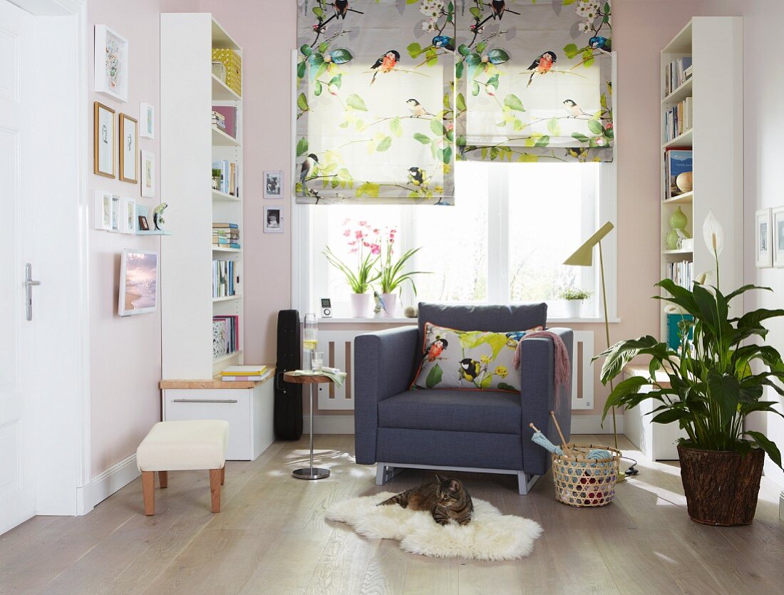 Sessel flankiert von weissen Bücherregalen und florale Faltrollos mit Vogelmotiv in sonniger Leseecke