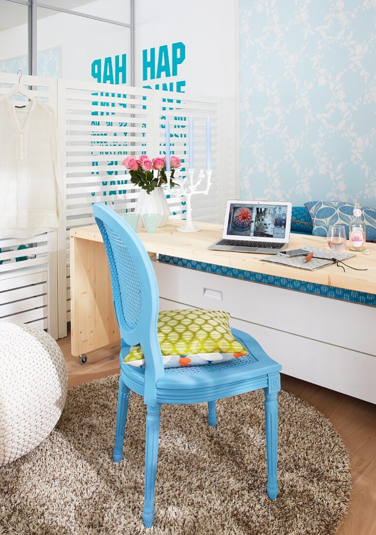 A wheeled wooden table used as a desk in front of a bed with a blue woven chair