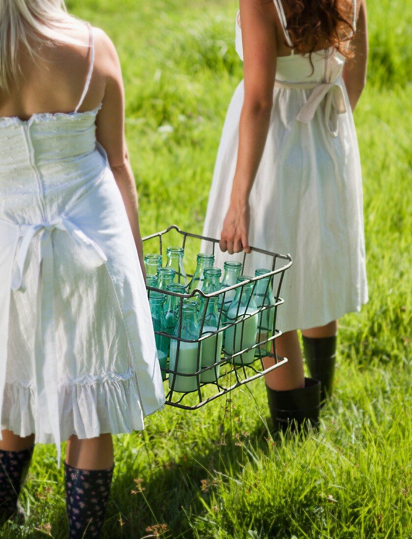 Junge Frauen in nostalgischen Sommerkleidern tragen mehrere Milchflaschen in einem Metallträger