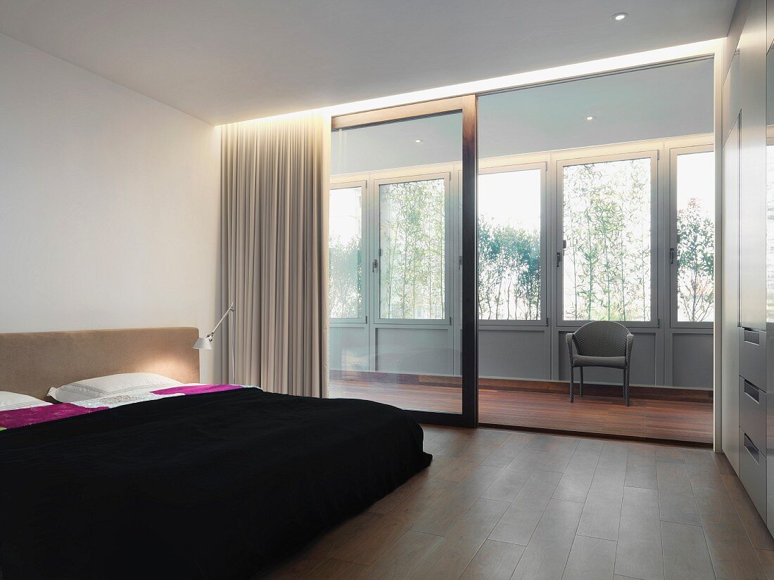 Doppelbett mit schwarzer Tagesdecke vor breiter Schiebetürfront zu verglaster Holzveranda