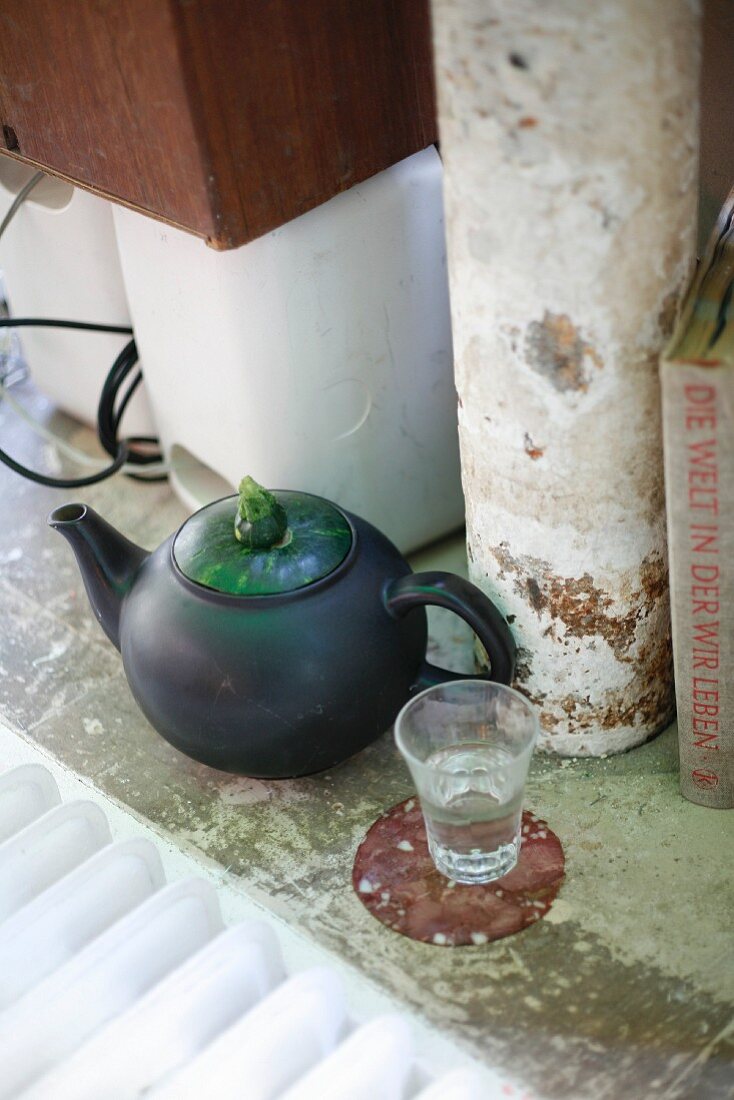 Schwarze Teekanne mit Zucchini-Deckel und Wasserglas auf Wurst-Untersetzer
