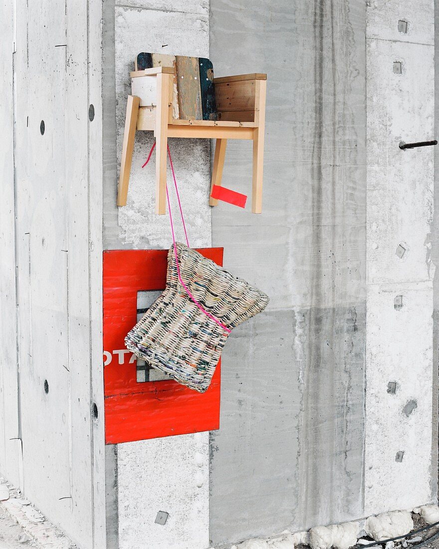 Hocker aus Zeitungspapier vor rotem Passepartout und Kinderstuhl aus Abrissholz an Betonwand aufgehängt