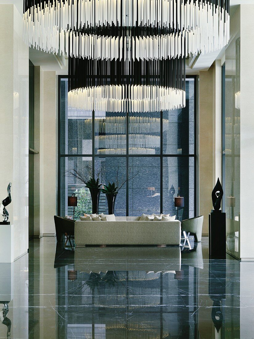 Empfangsraum mit ringförmigen Hängelampen aus leuchtenden Stäben und weisser Sofagarnitur vor Fensterfront auf spiegelndem Steinboden