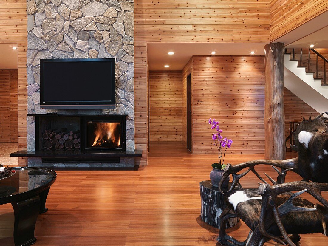 Minimalistischer Wohnraum mit holzverkleideten Wänden und rustikaler Stuhl mit Fellsitzfläche vor offenem Kamin unter Fernseher an Natursteinwand