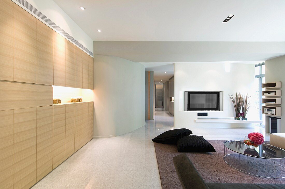 Einbauschrank aus hellem Holz mit indirekter Beleuchtung in offenem Wohnraum und Blick auf Fernseher im Raumteiler