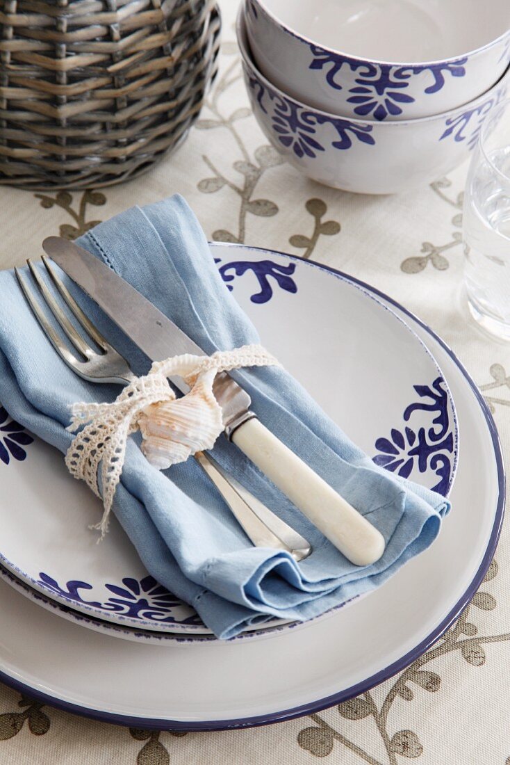 Place setting with blue-patterned china; old silverware and shell tied to pale blue napkin