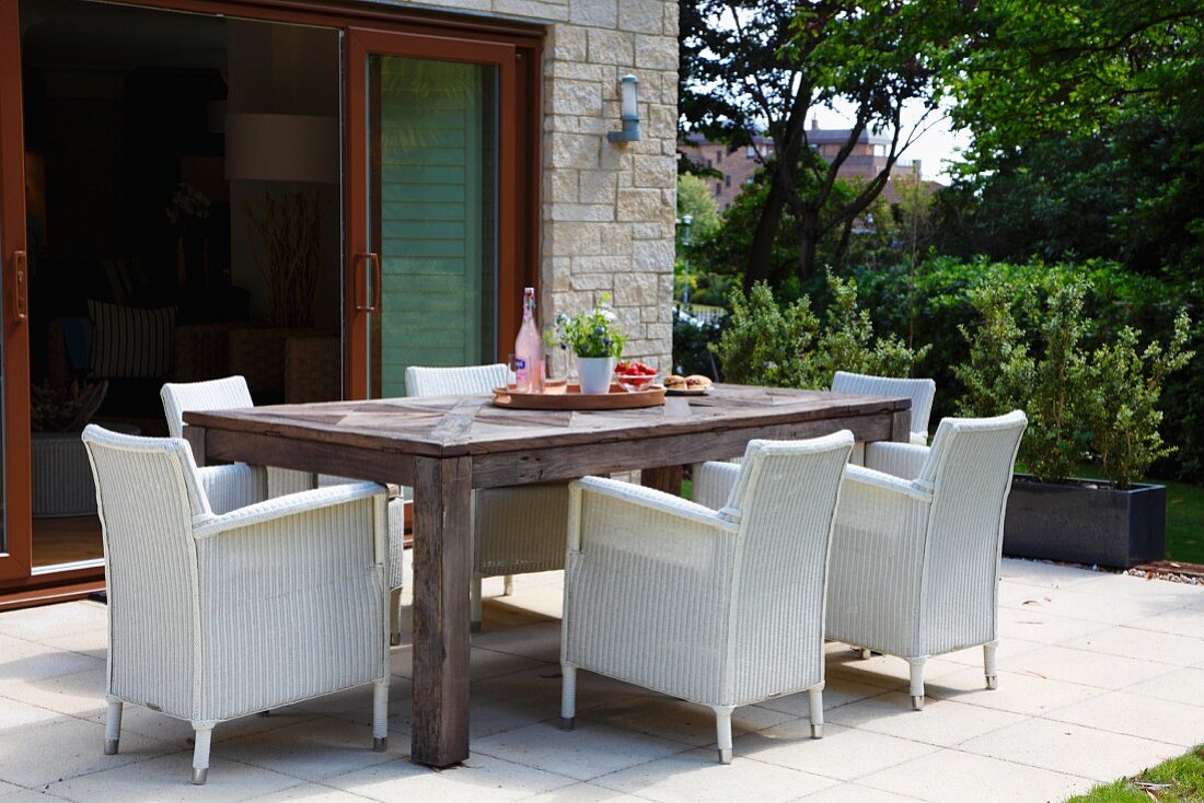 White wicker armchairs around large wooden table in front of open terrace doors in garden