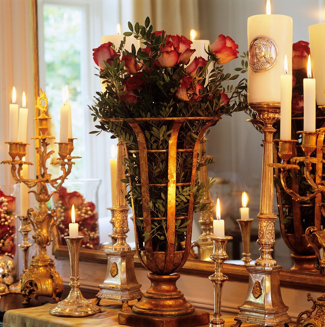 Rosenstrauss in einer Messing Vase umringt von silbernen Kerzenständern mit brennenden Kerzen vor Wandspiegel