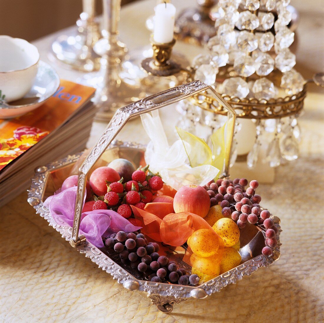 A silver basket of artificial fruits in front of a table lamp with crystal decoration