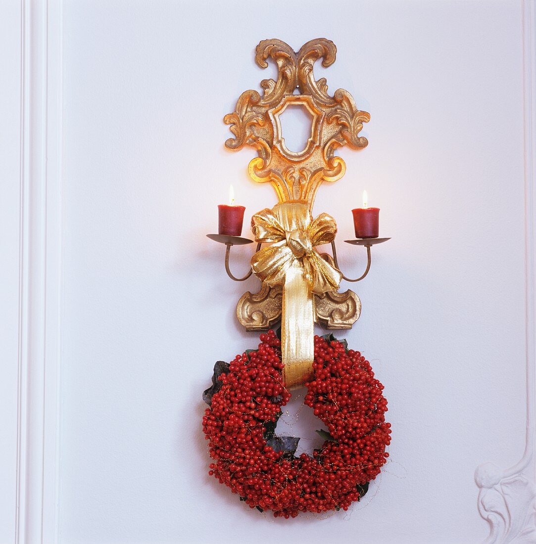 Gilt wall bracket with lit candles and wreath of red berries on white wooden wall