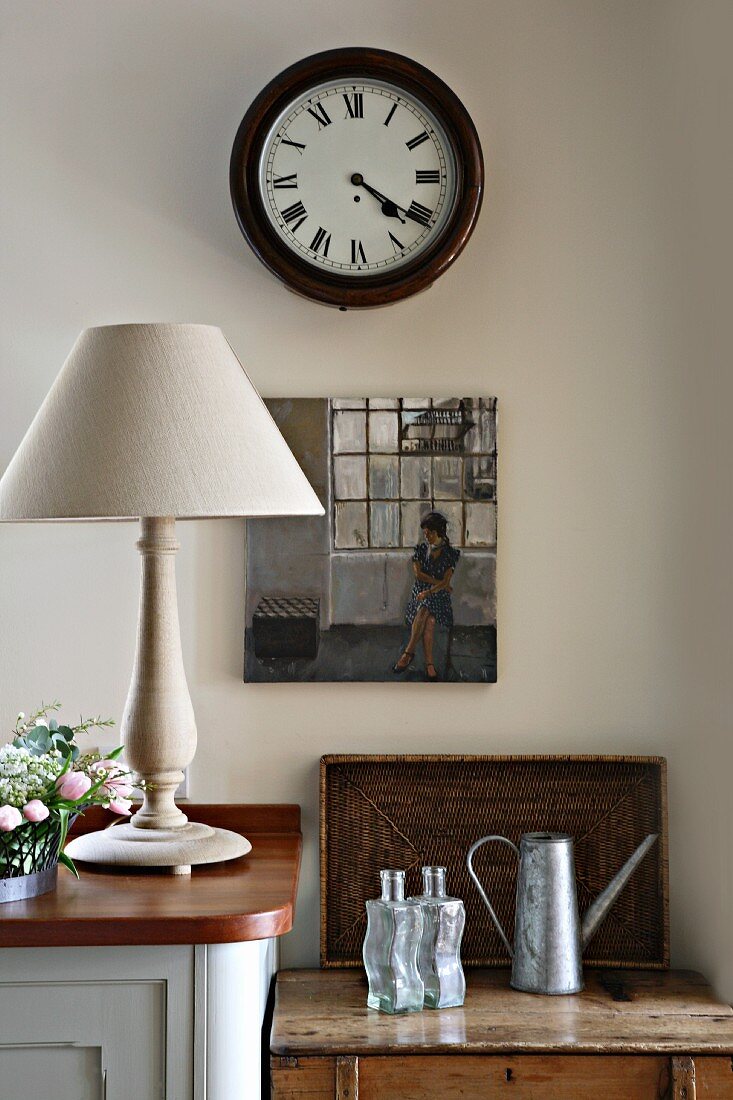 Wall clock with Roman numerals and small oil painting above tin watering can, wooden tray and glass bottles on wooden trunk lid next to table lamp with turned base on wooden sideboard