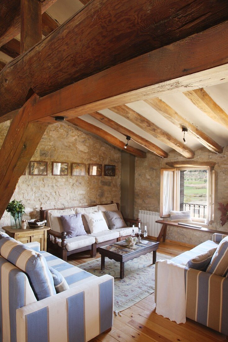 Seating area with striped sofas in tasteful country house style below rustic roof beams in historical building