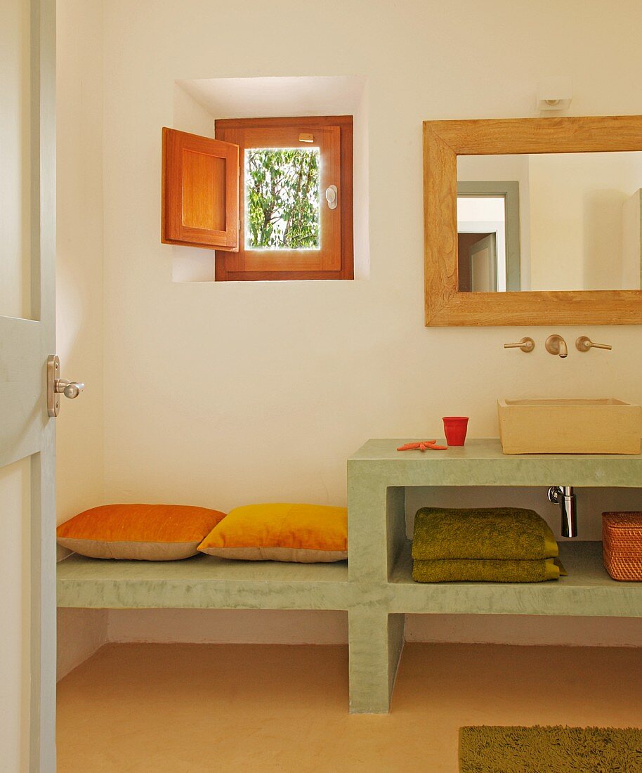 View through open door of simple, concrete washstand with integrated bench against wall below small window with wooden shutter