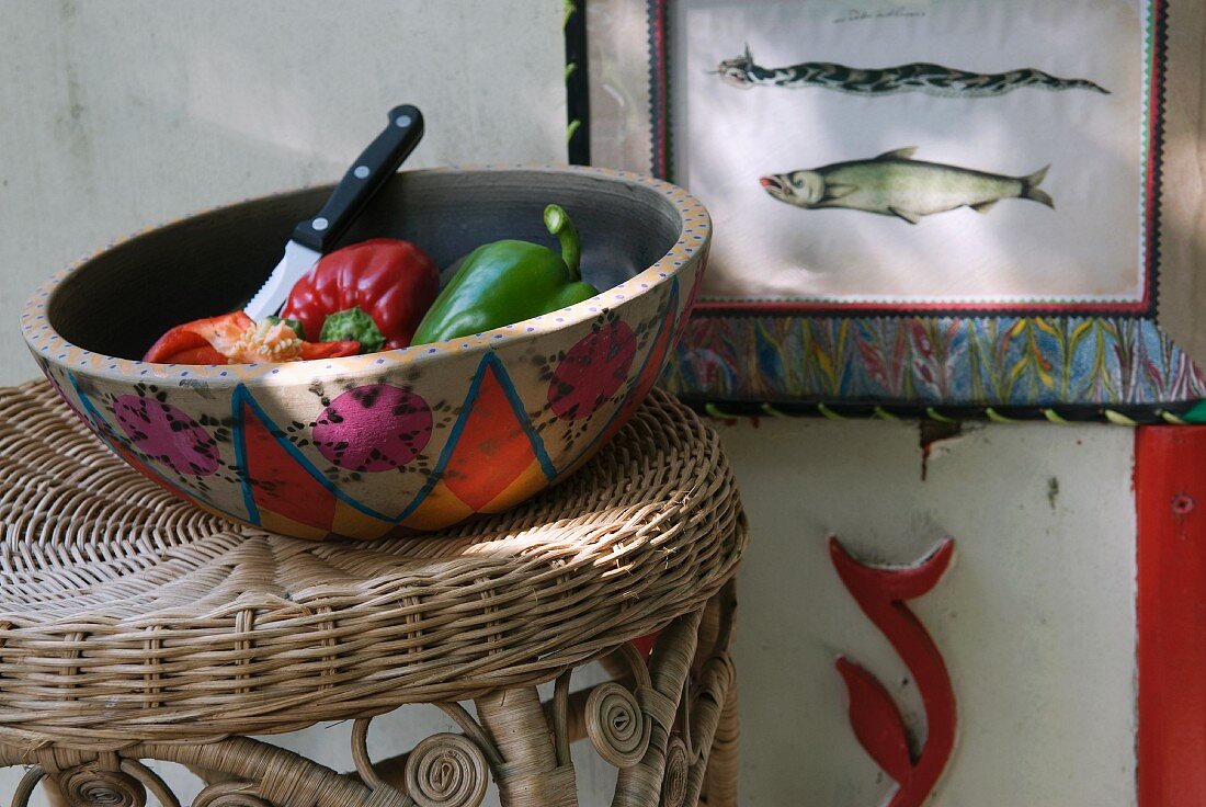 Bunte Keramikschale mit Gemüse auf geflochtenem Hocker; Fischdarstellungen auf gerahmtem Bild an der Wand