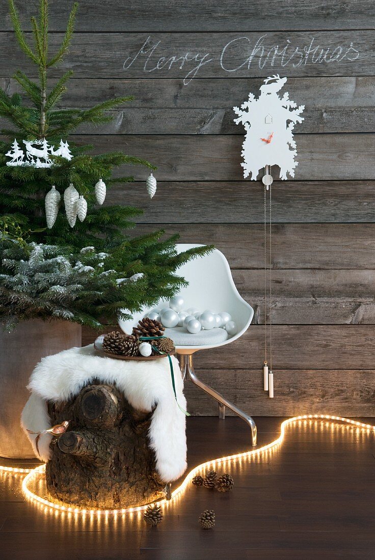 Lichterkette schlingt sich um Baumstumpf mit Fell und geschmücktem Weihnachtsbaum vor Vintage Holzwand mit Kuckucksuhr