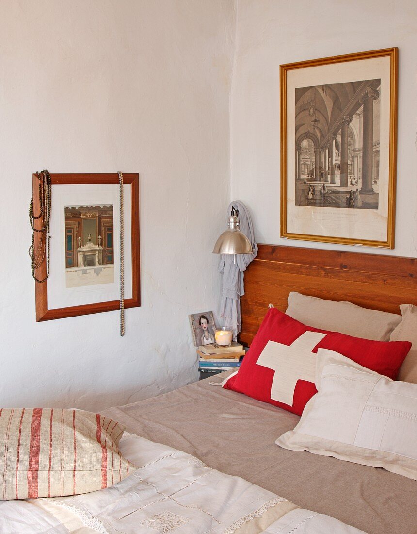 Rotes Kissen mit weißem Kreuz auf schlichtem Bett in Zimmerecke und gerahmte Photos an Wand