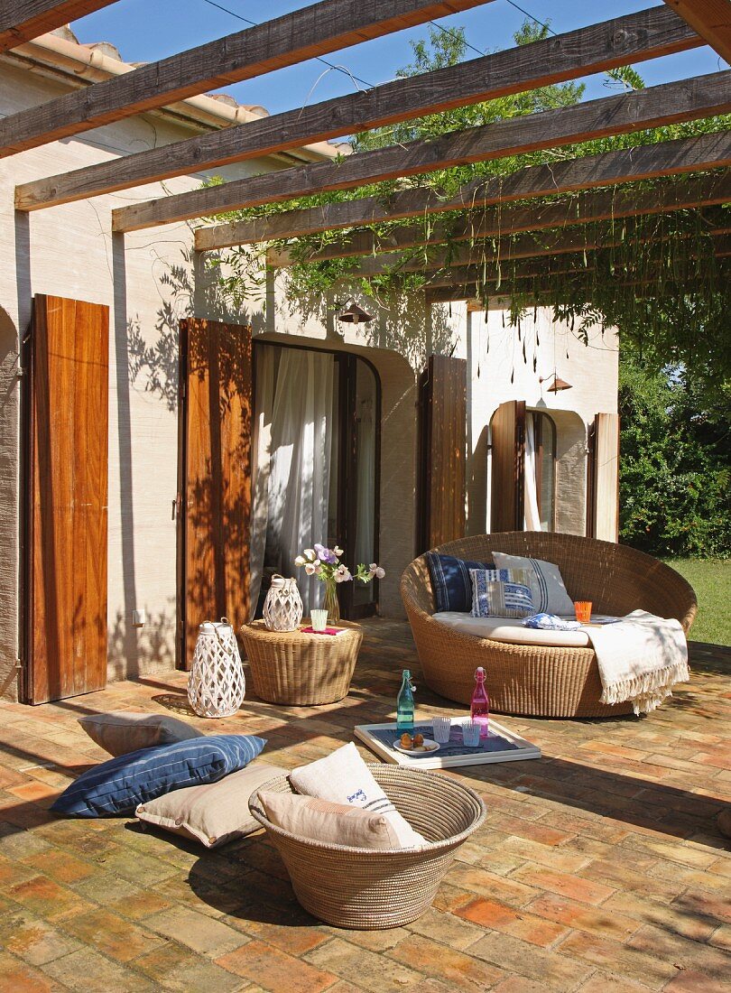 Sonnenbeschienene Terrasse mit Rattan Loungesessel und Kissen unter Pergola vor mediterranem Haus