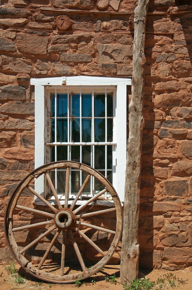 Vergittertes Fenster eines Steinhauses; davor ein antikes Wagenrad