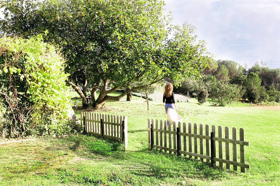 Green, summer landscape with picket fence and woman walking across field beyond