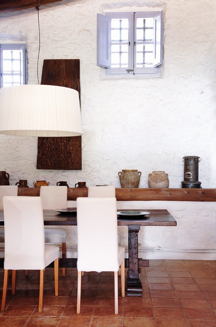 Essbereich mit massivem Tisch und gepolsterten weißen Stühlen, dahinter Wandboard mit Krugsammlung und Tontöpfen an weiß getünchter Wand mit Sprossenfenstern in der Dachschräge