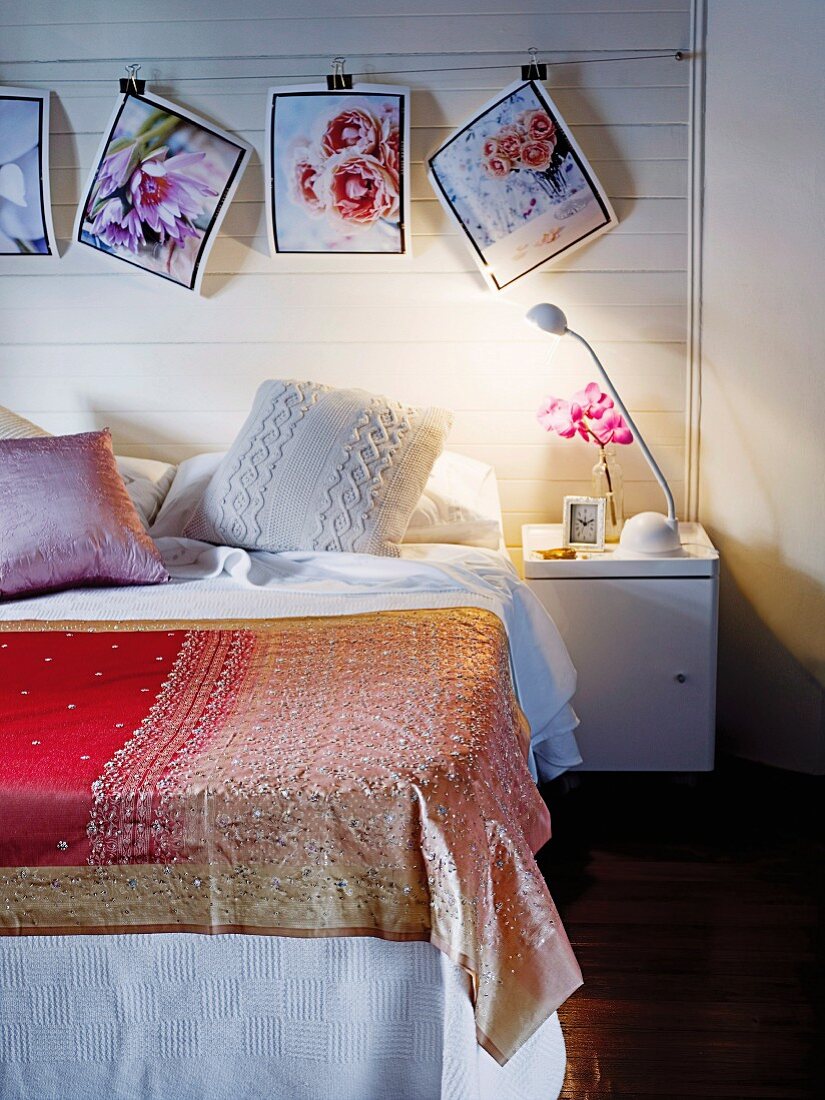 Bedspread on simple bed below floral photos hanging from clips on white wooden wall