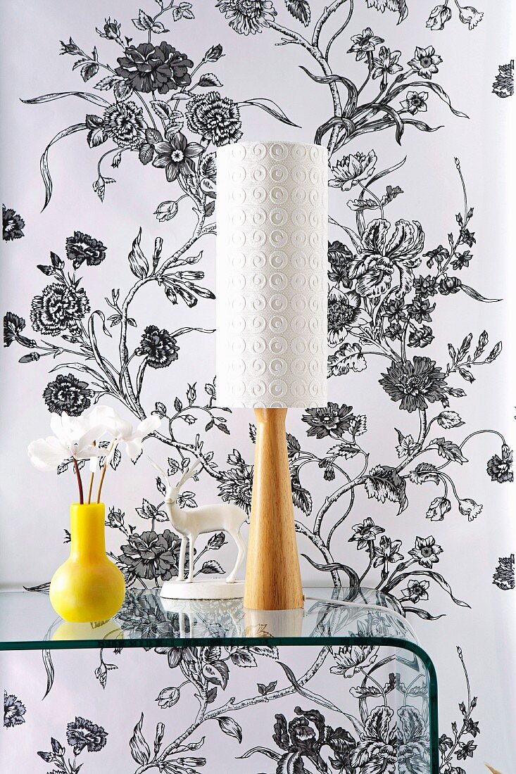 Gelbe Vase neben Tischlampe mit weißem Schirm und Fuss aus Holz auf Glas Konsolentisch-rundgezogen vor tapezierter Wand mit schwarz-weißem Blumenmuster
