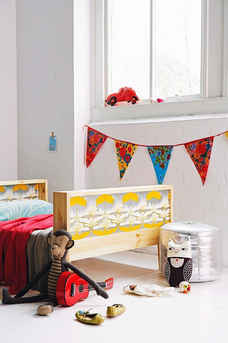 Boxy, wooden, DIY child's bed with painted headboard and foot and toys on floor in front of window with string of bunting