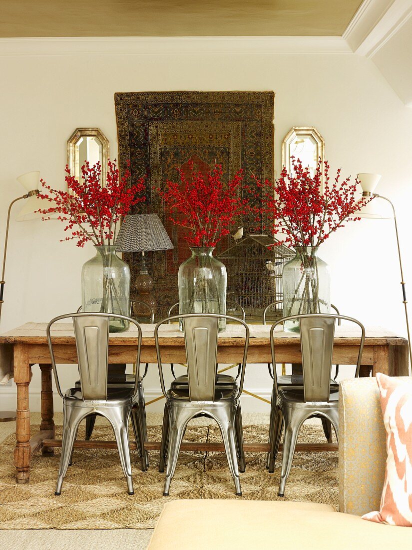 Retro metal chairs around wooden table with branches of berries in vases in front of tapestry hanging on wall