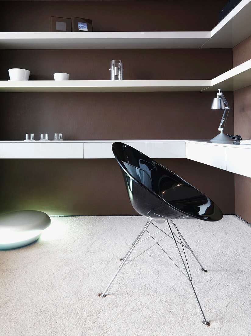 Black shell chair in front of wall mounted shelving on a brown wall in a living room corner