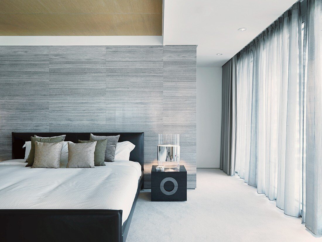 Elegantes Designer Schlafzimmer mit Doppelbett vor gefliestem Raumteiler in Grau neben bodenlangen Vorhängen am Fenster