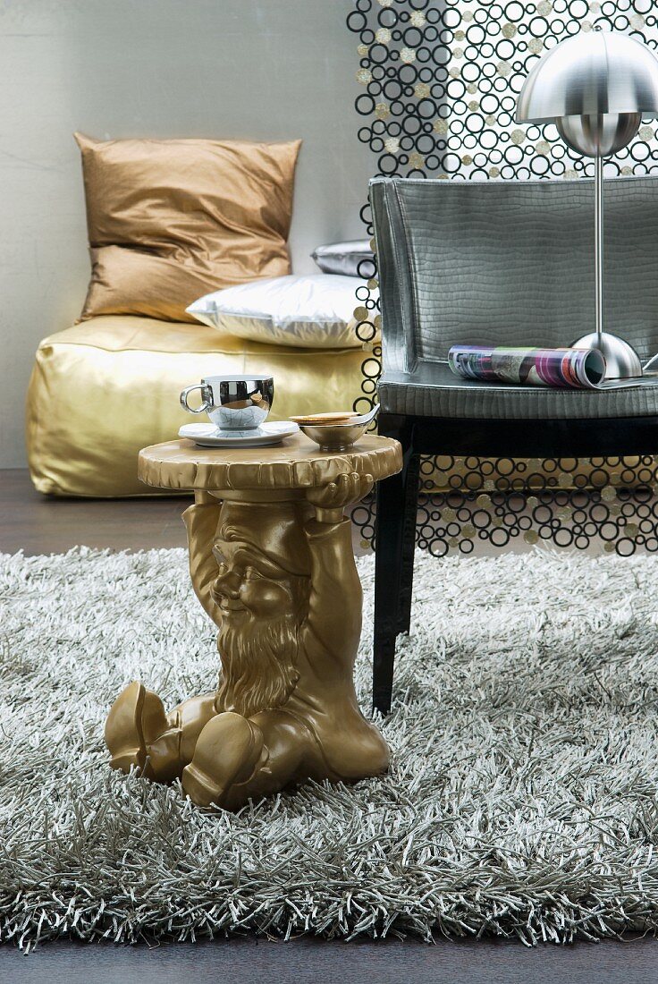 Goldfarbener Beistelltisch mit Gartenzwergfigur als Fuss und Stuhl auf Flokatiläufer vor goldfarbenem Polster