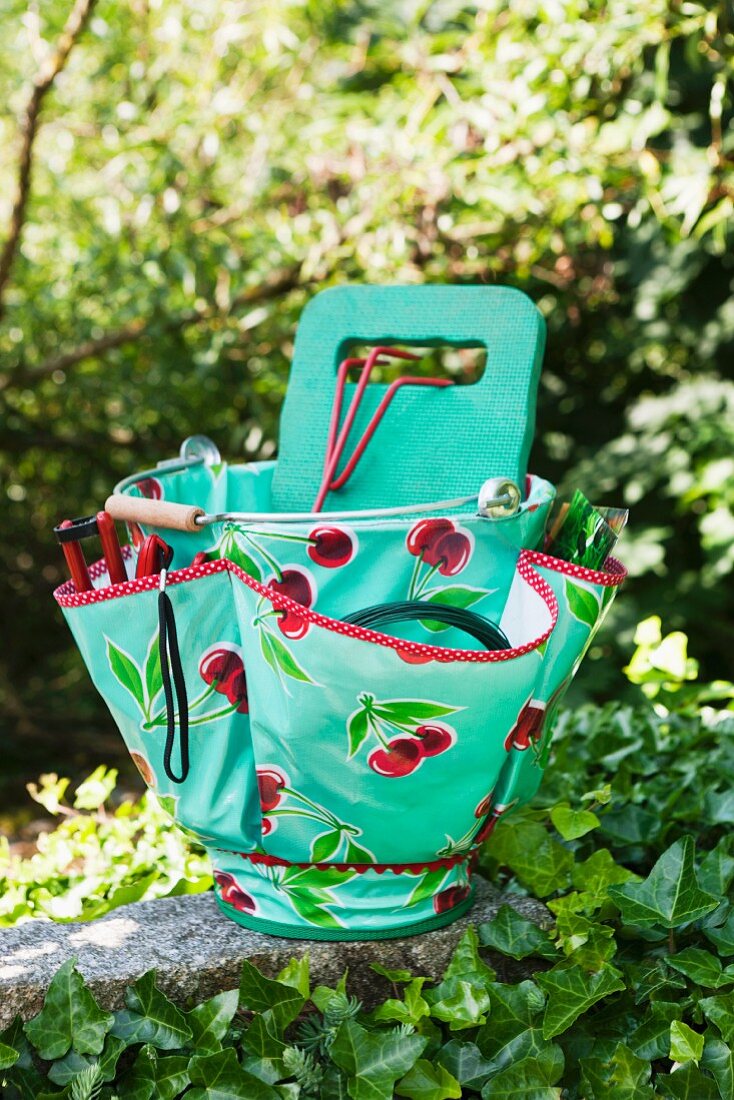 Gartengeräte in türkisfarbener Plastiktasche mit Kirschen Motiven auf Steinmauer im Garten