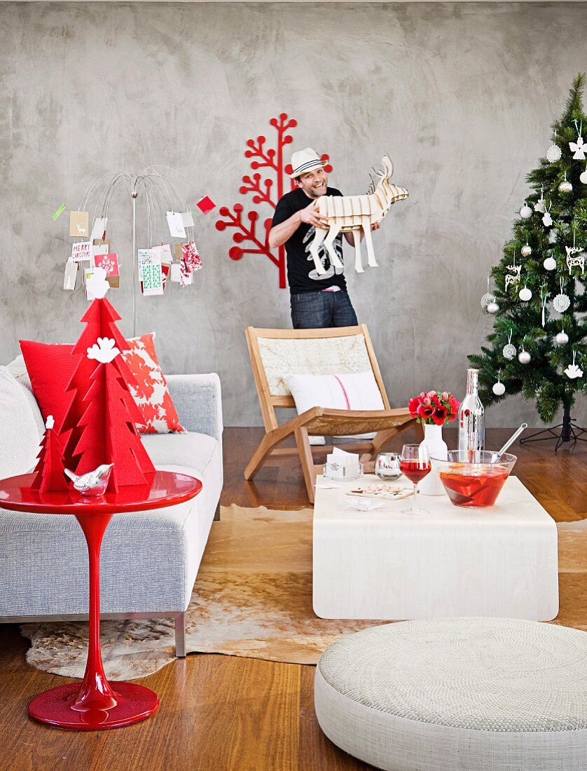 Christmas atmosphere - Christmas decorations on red side table in front of designer sofa and coffee table and man holding reindeer ornament next to Christmas tree